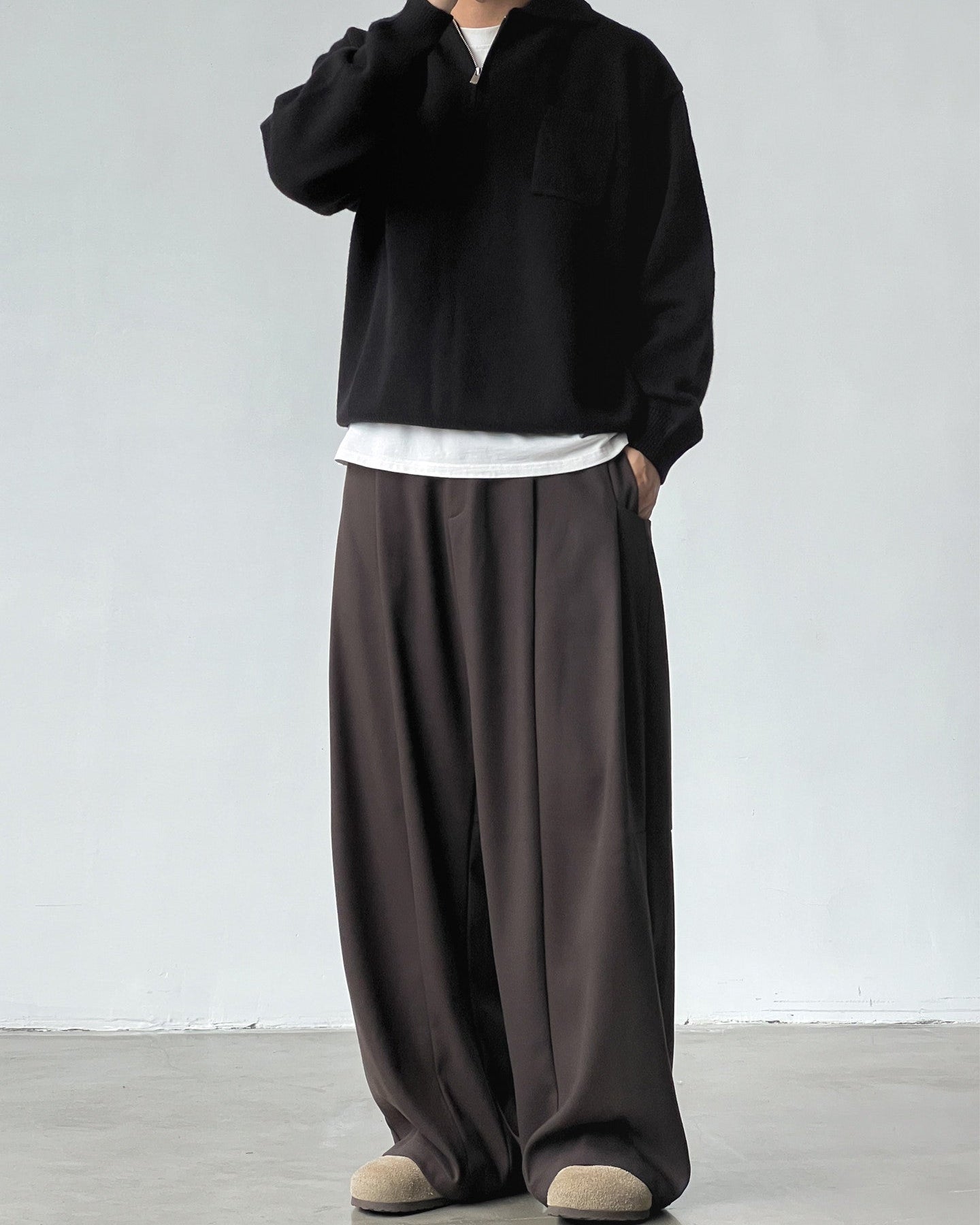 Person wearing a black sweater, white shirt, and brown wide-leg pants against a plain background